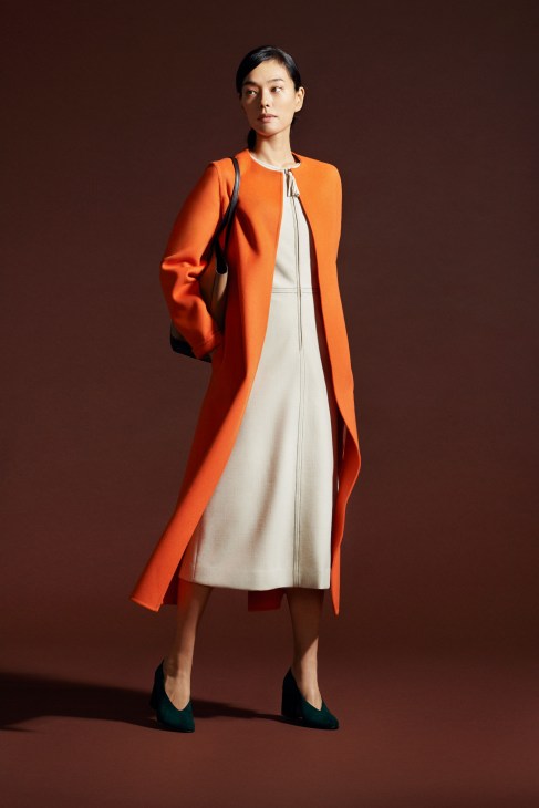 Woman in light camel coat with black dress and black shoes carrying black bag standing against white background