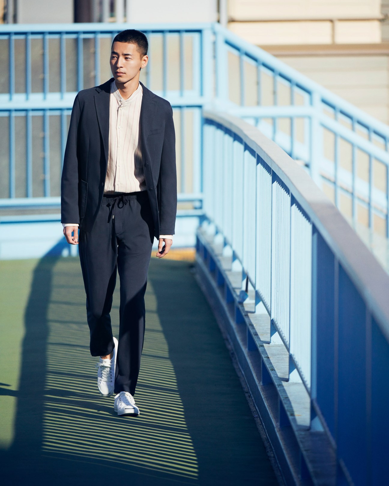 Man in black Circolo 1901 jacket and trousers with Pal Zileri shirt and Converse trainers on a blue pedestrian bridge.