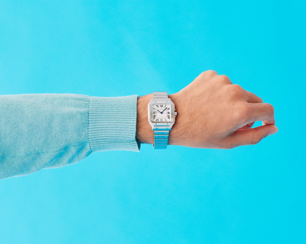 Man's wrist wearing a Cartier watch with a turquoise jumper sleeve against a bright blue background.