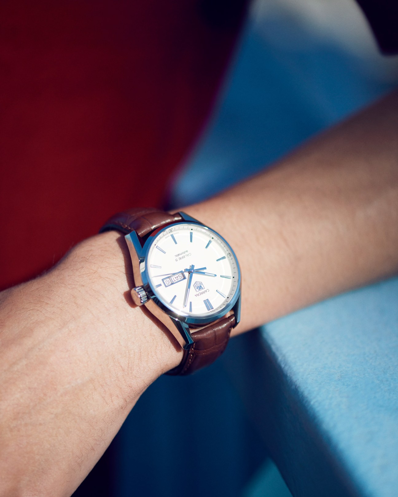 Man's wrist wearing Tag Heuer watch with brown leather strap and white dial with blue accents.
