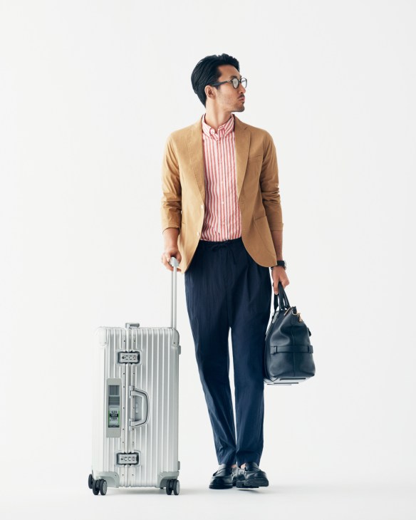 Man wearing camel blazer and striped shirt with navy trousers, holding Rimowa suitcase and Bally bag.