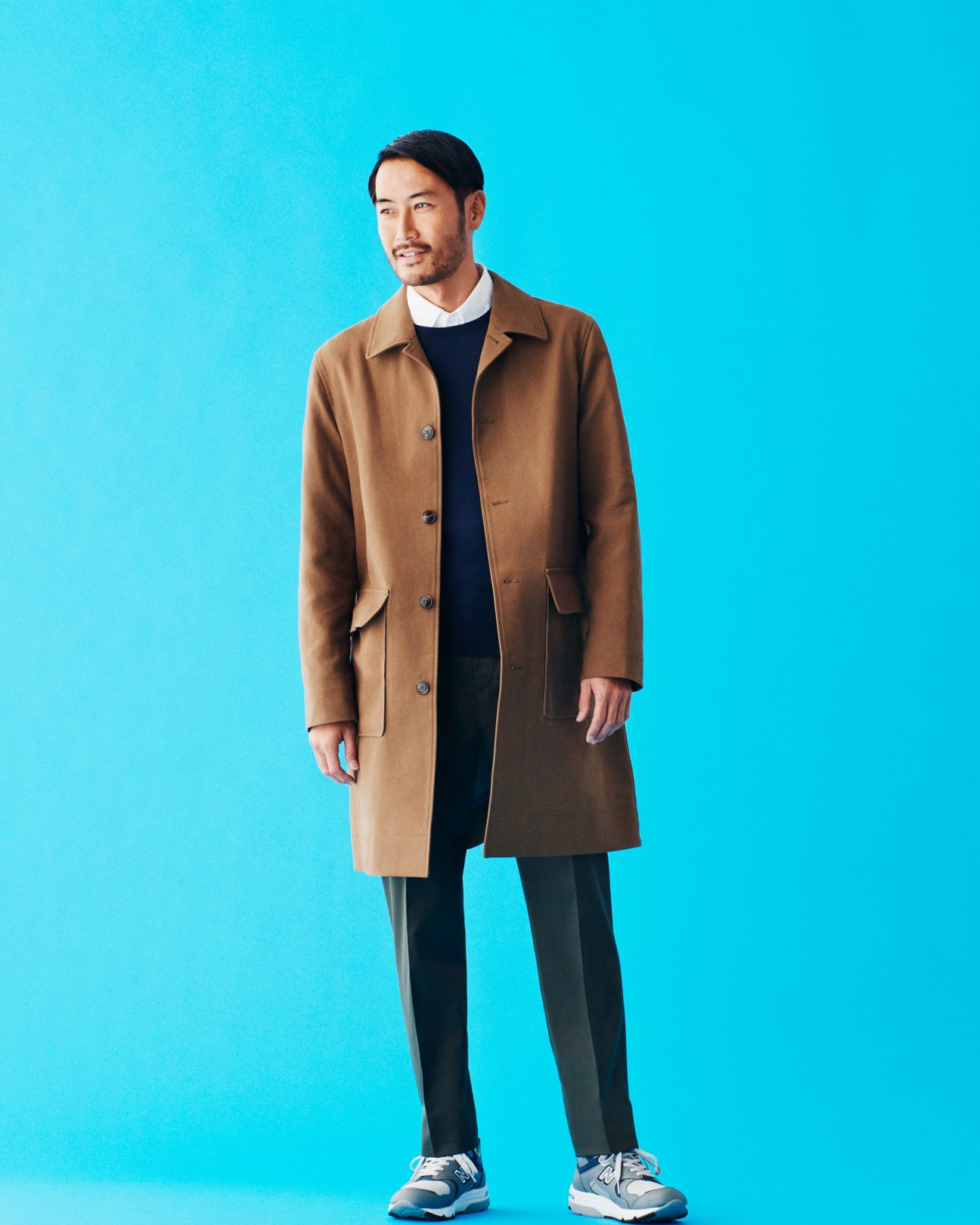 Man wearing a camel coat, navy jumper, white shirt collar, dark trousers, and navy trainers against a bright blue background.