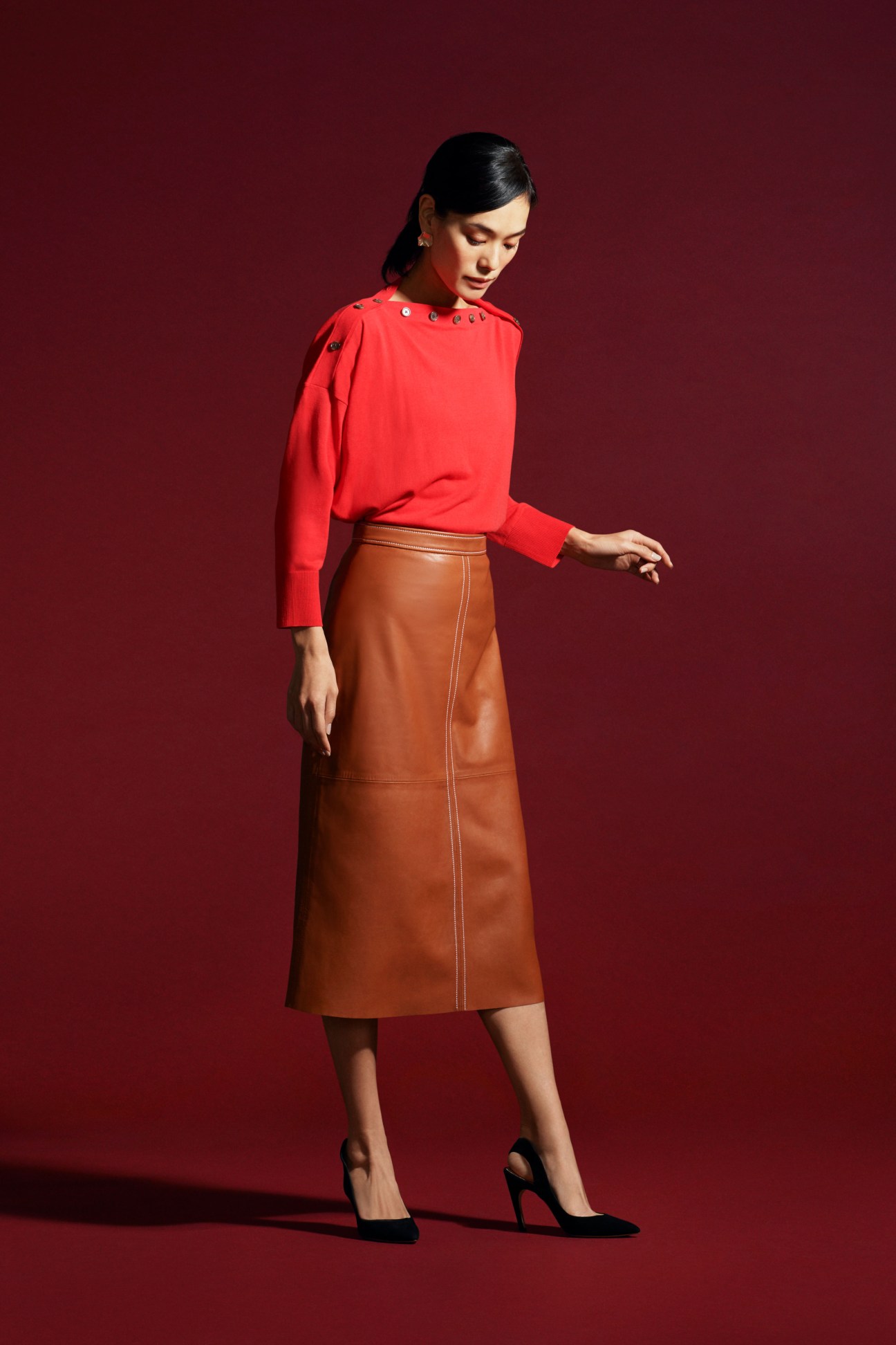 Woman in cream sleeveless top with brown leather skirt and black heels against red background