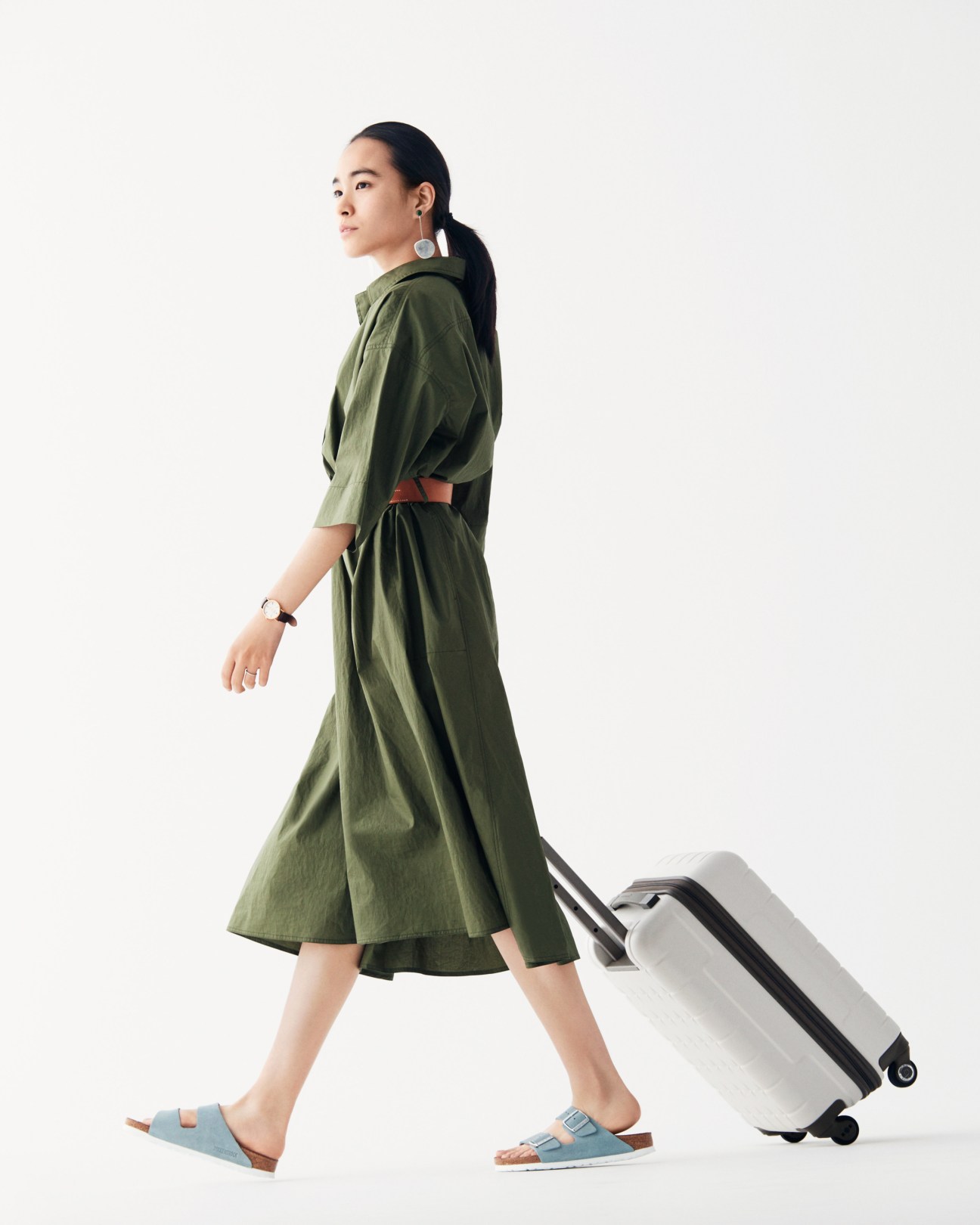 Woman in olive green dress with Hermès belt and Birkenstock sandals pulling a white Proteca suitcase.
