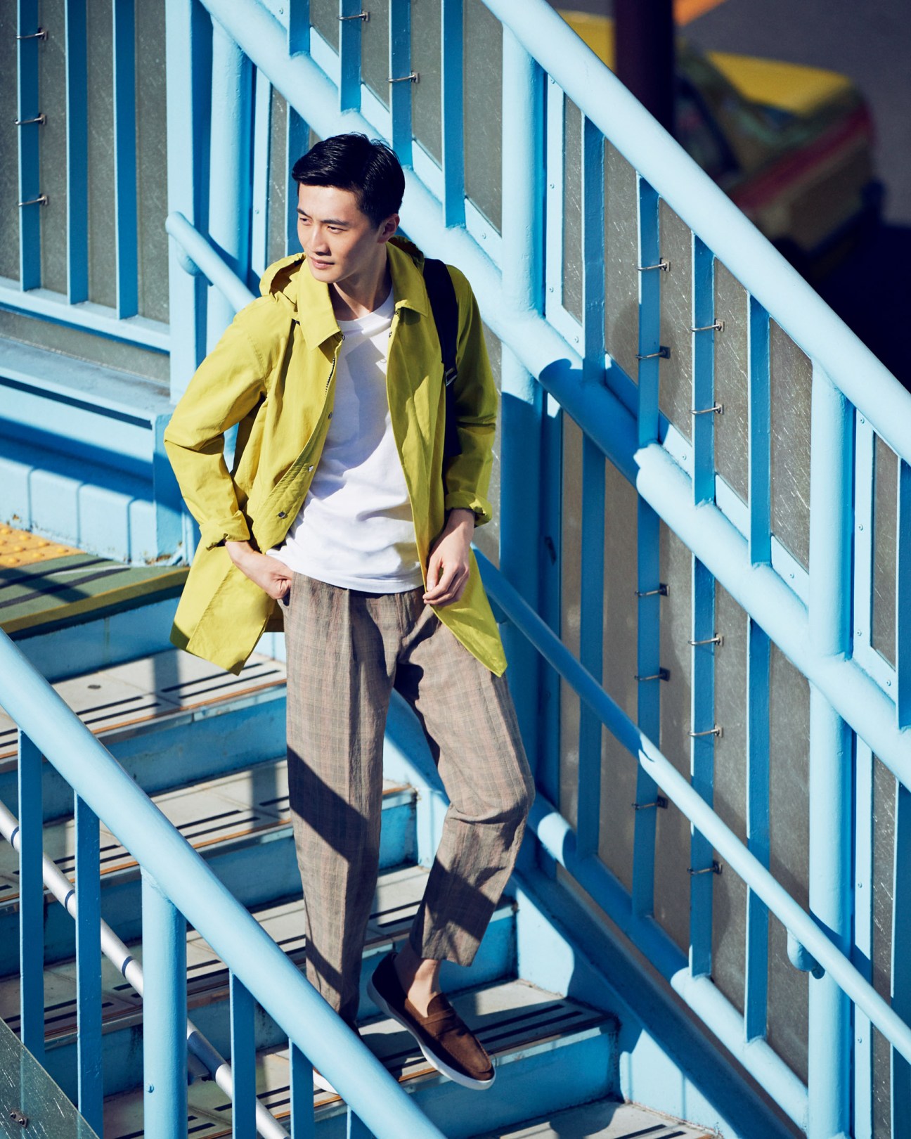 Man on steel-and-concrete pedestrian bridge wearing yellow coat, white t-shirt, and neutral trousers.