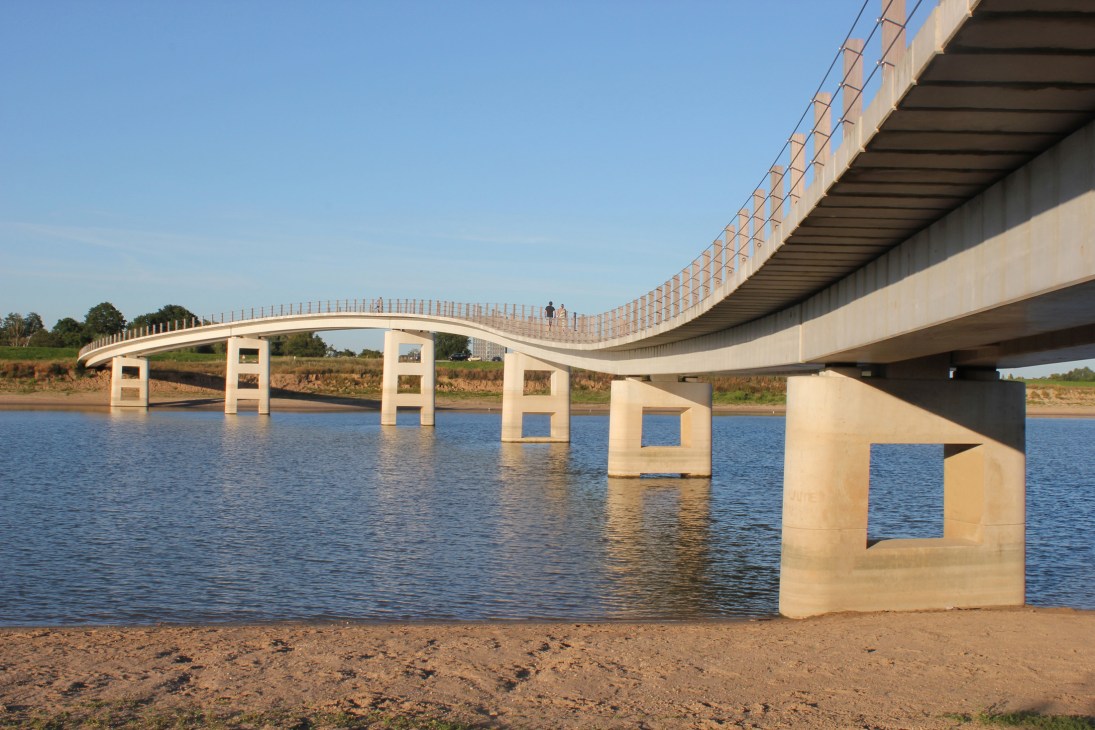The Zalige Bridge's undulating concrete structure spans across water with tilted support columns and a curved pedestrian walkway.