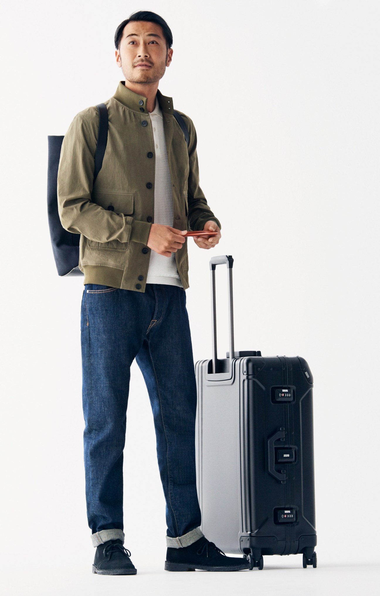 Man in tan Valstar jacket, Altea polo shirt, and Brooksfield jeans holding passport, standing with Zero Halliburton suitcase.