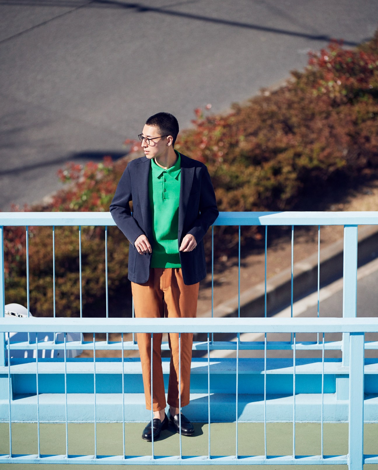 Model wearing navy Eleventy jacket, green Sloane polo shirt, orange Tomorrowland trousers, and Crockett & Jones shoes on a blue pedestrian bridge.