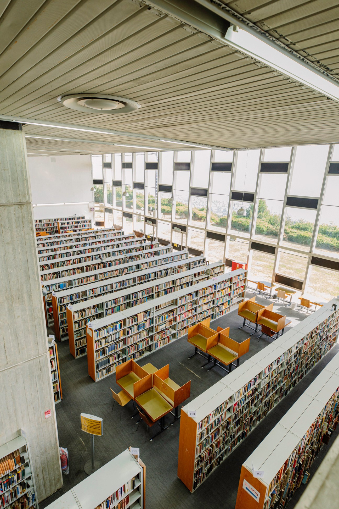 Interior of University of Haifa library with stepped bookshelves, yellow chairs, and floor-to-ceiling windows overlooking landscape.