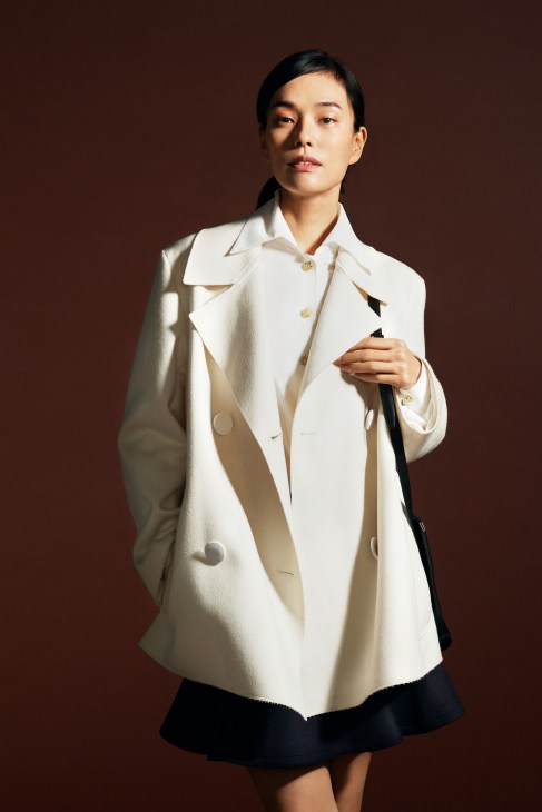 Woman in beige coat with white shirt and dark skirt carrying brown bag standing against white background
