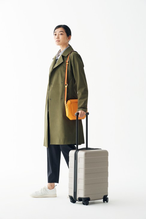 Woman in olive coat with orange shoulder bag holding beige suitcase for travel.