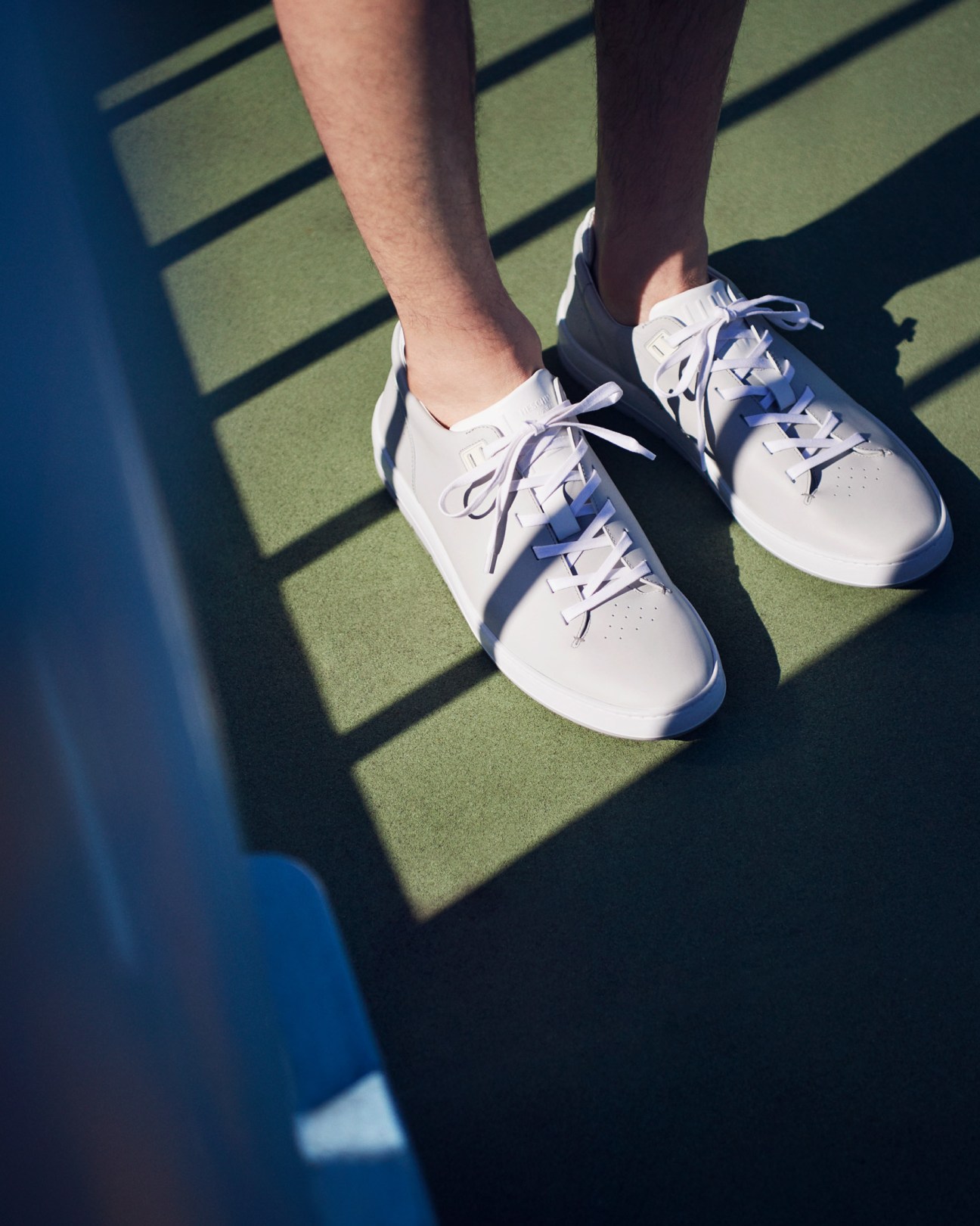 White leather trainers with blue laces on feet, cast shadow on green pedestrian bridge surface.