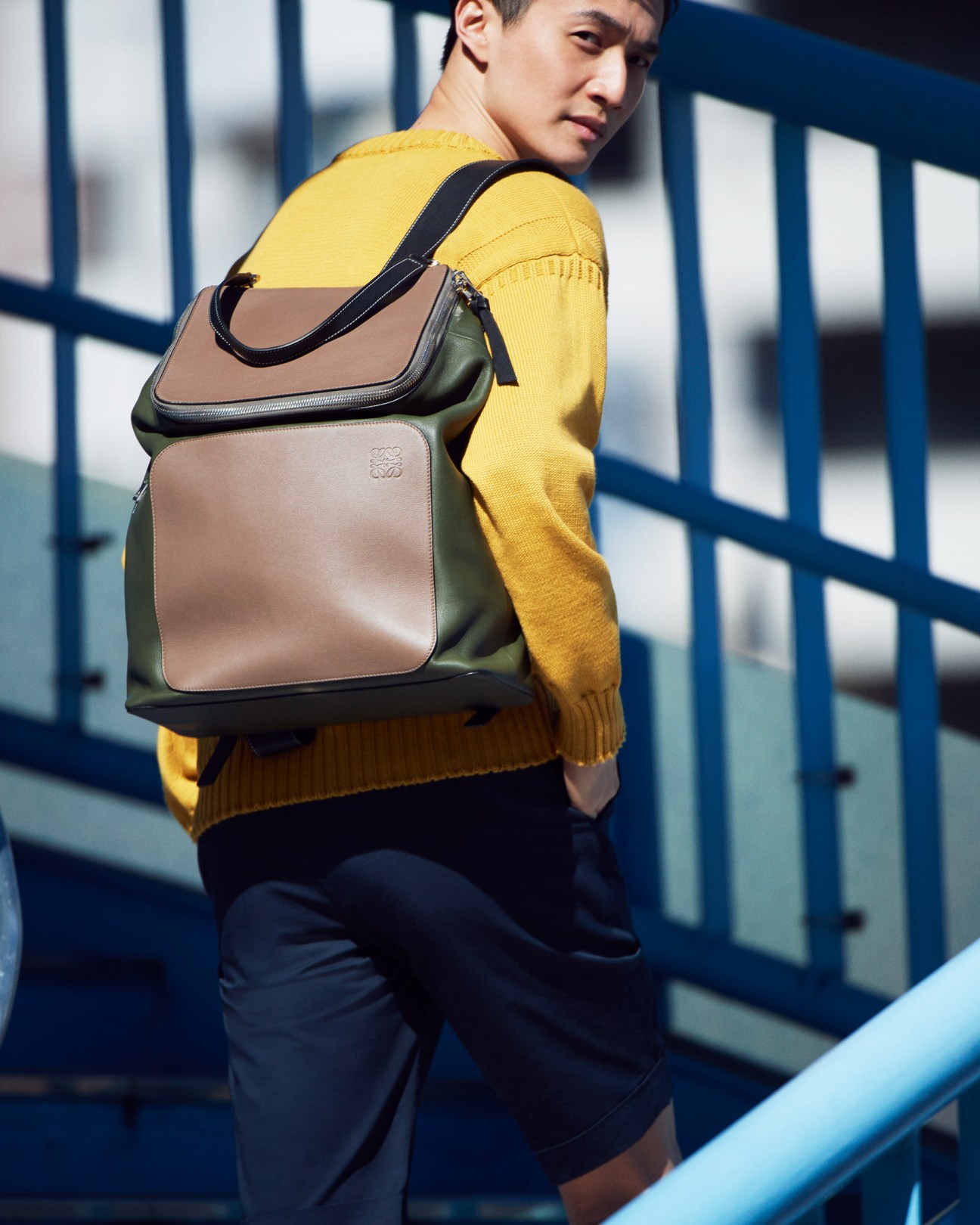 Model wearing yellow jumper and navy shorts with tan and olive Loewe backpack on blue pedestrian bridge.