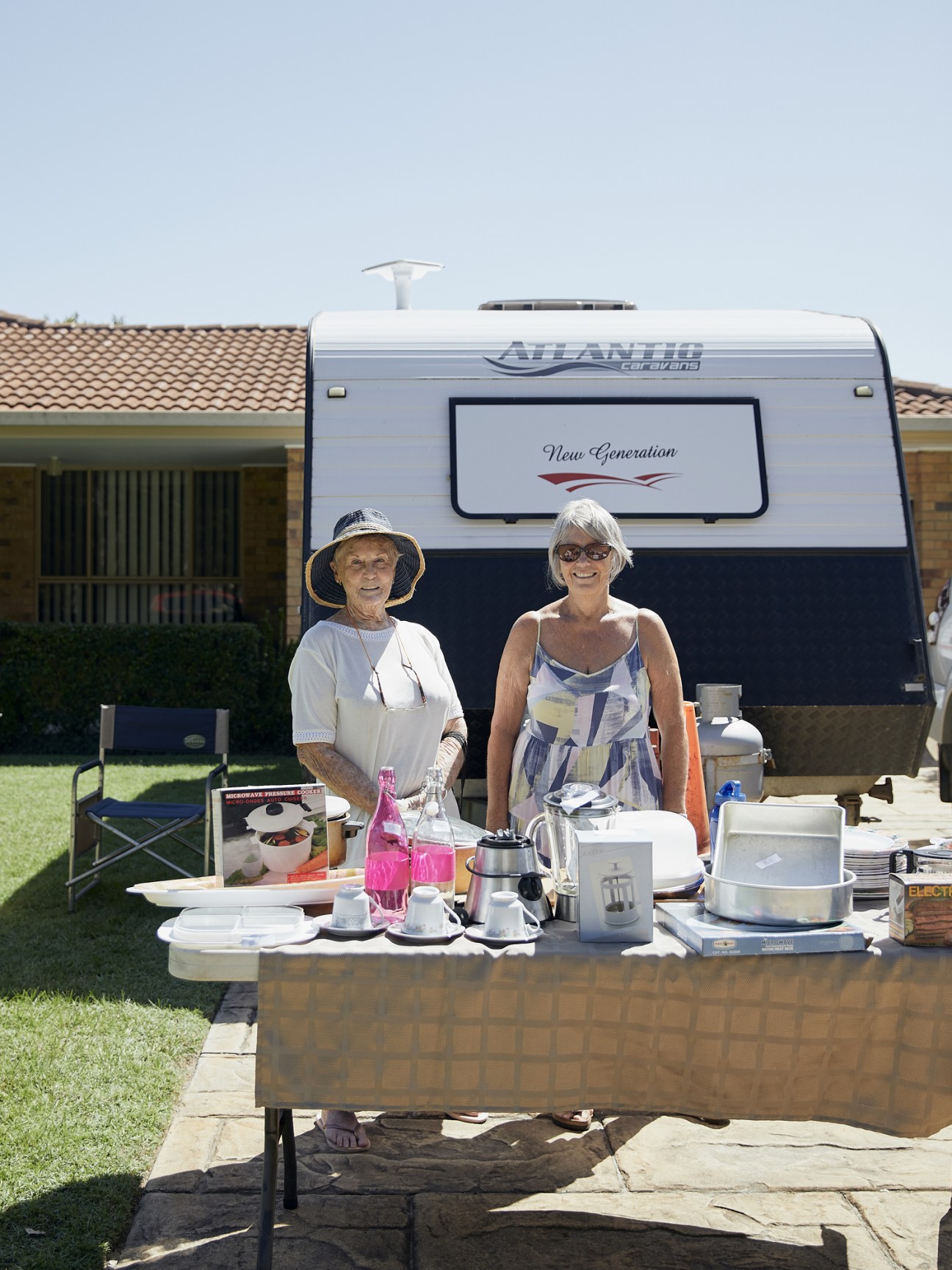 Two older adults stand at a table with kitchen items and beverages in front of an Atlantis caravan parked on a lawn.