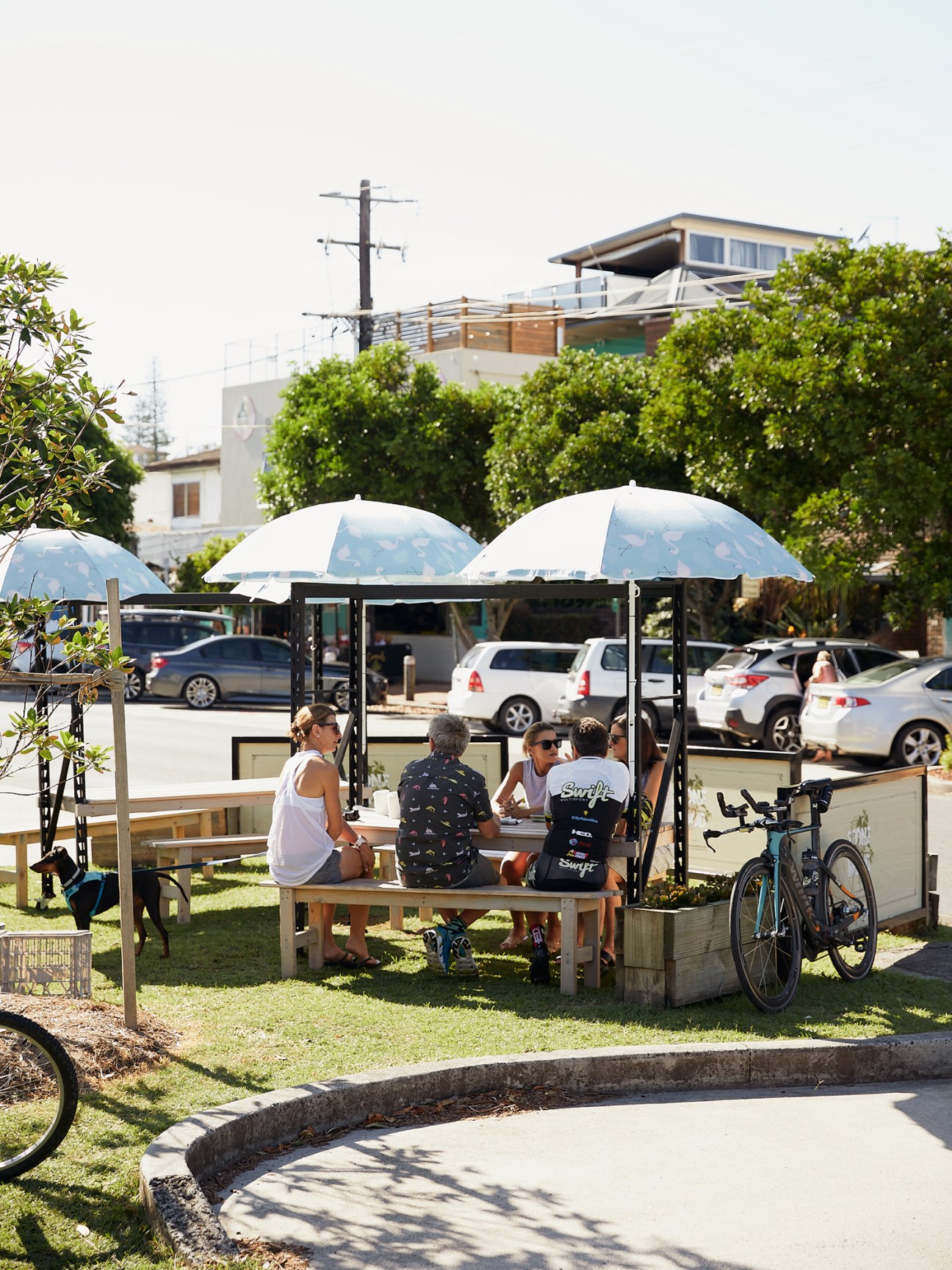 People seated under white umbrellas at a covered outdoor dining area near a parking lot and buildings in Yamba.