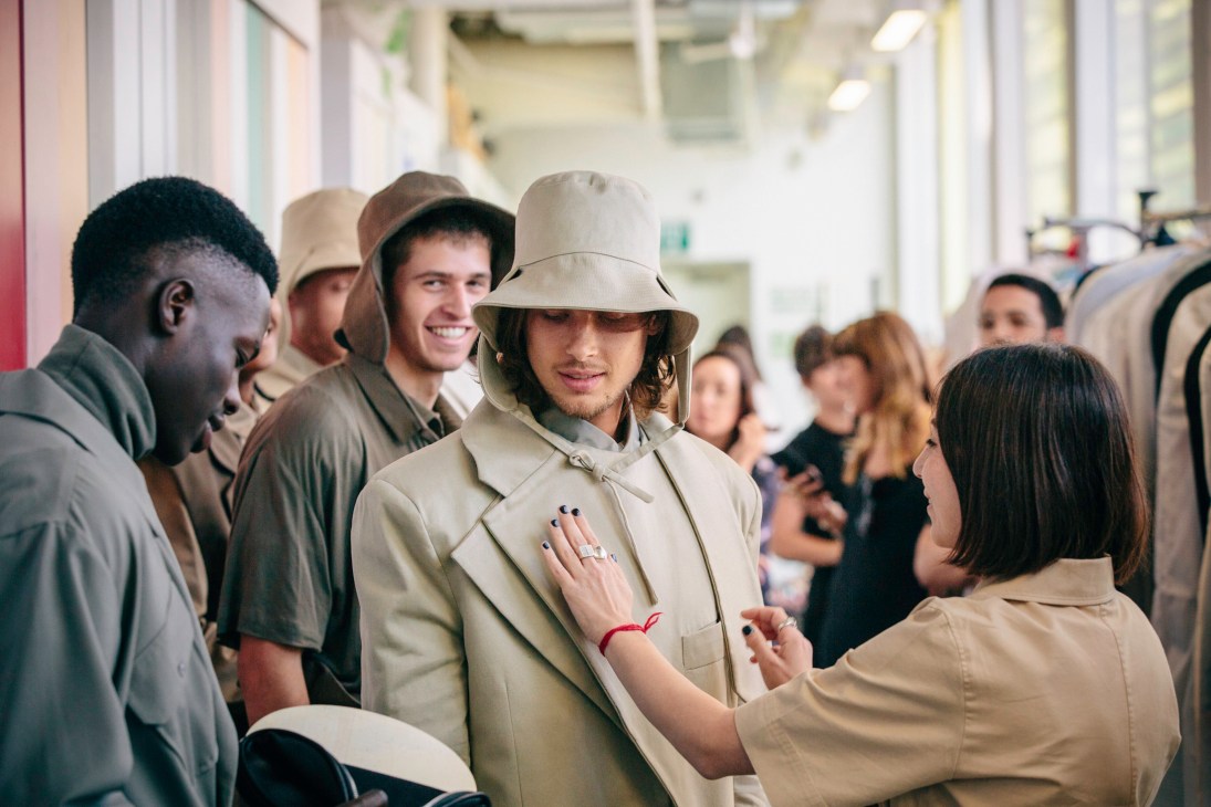 A model wearing a beige button-up shirt and matching bucket hat stands in a backstage hallway surrounded by students and staff during Shenkar College's graduation runway show.