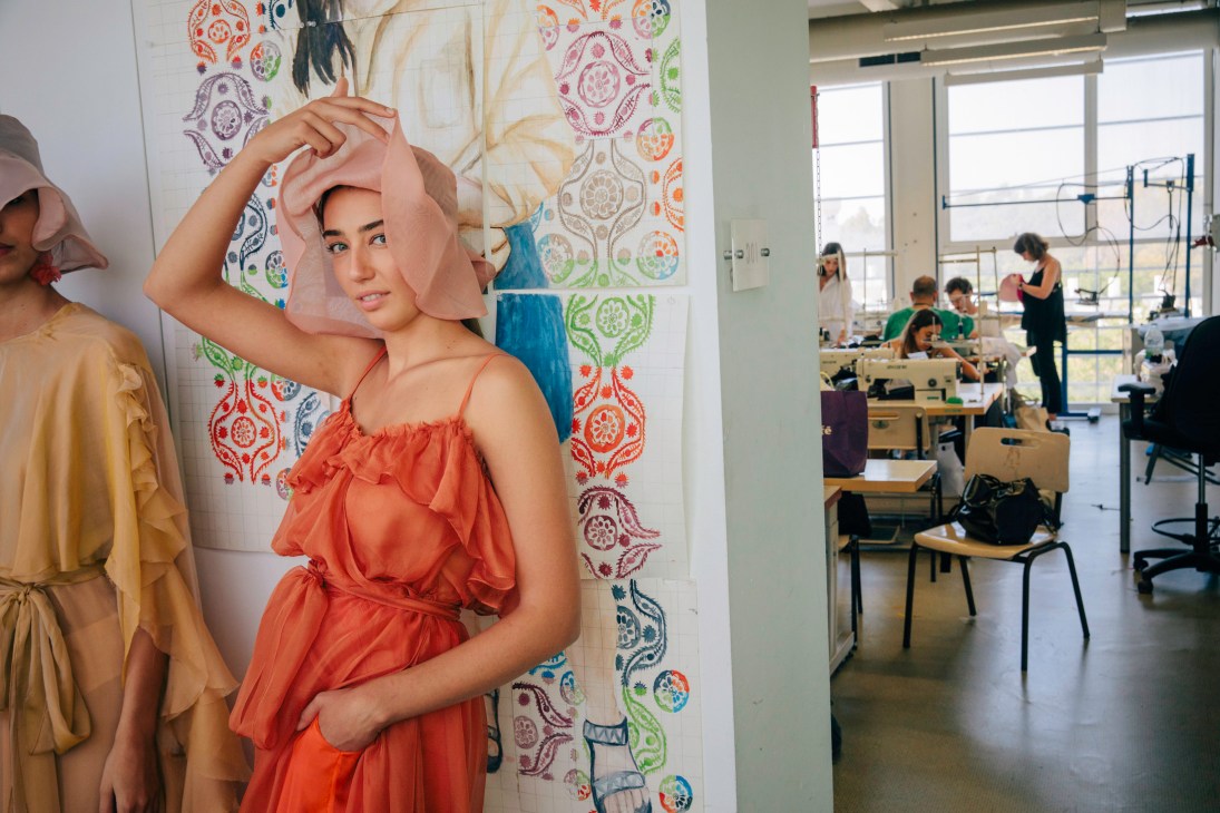 A mannequin wearing an orange ruffle dress and sheer fabric head piece stands in a classroom with patterned fabric samples on the wall behind it.