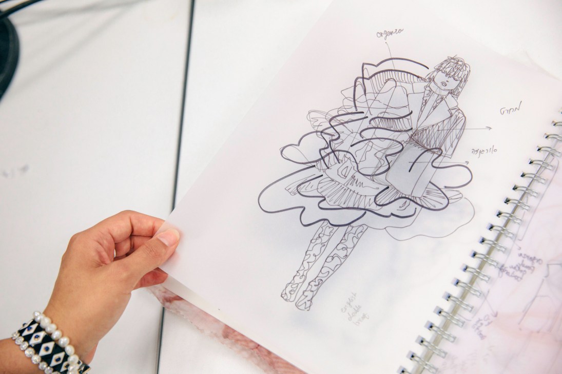 A student's hand holding a design sketchbook with pen drawings of fashion figures wearing layered, voluminous garments.