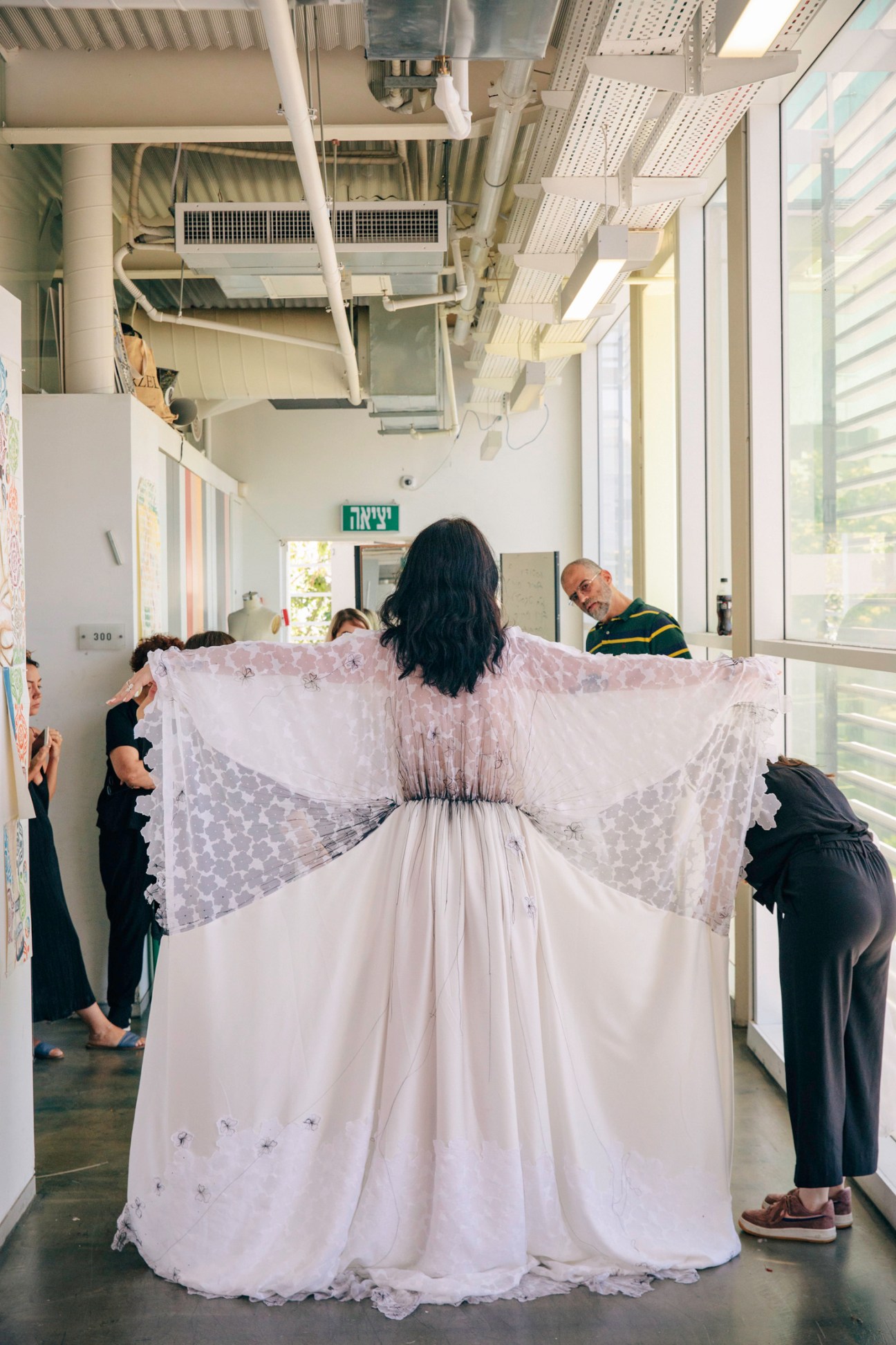 A model wearing a white lace dress with dramatic wing-like sleeves being fitted by instructors in a Shenkar College studio.