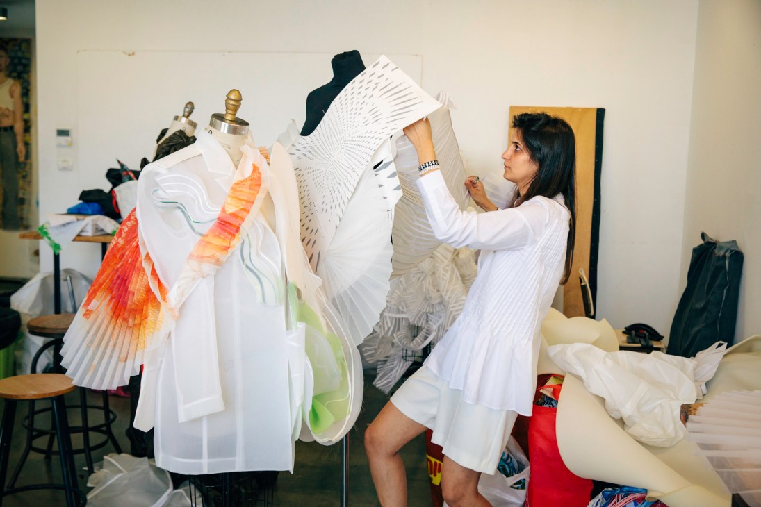 Student Shir Naeh adjusting white pleated fabric on a dress form in a fashion studio.