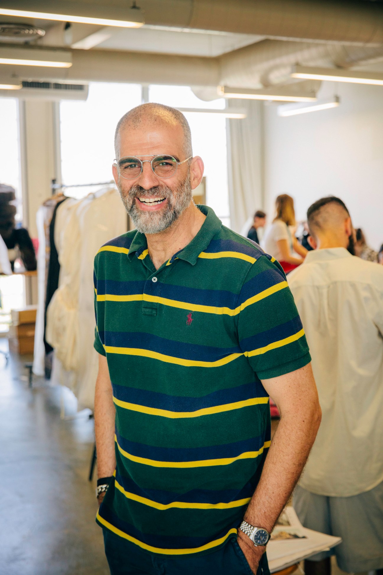 Professor Ronen Levin smiling in the Shenkar fashion department studio with students and clothing racks in background.