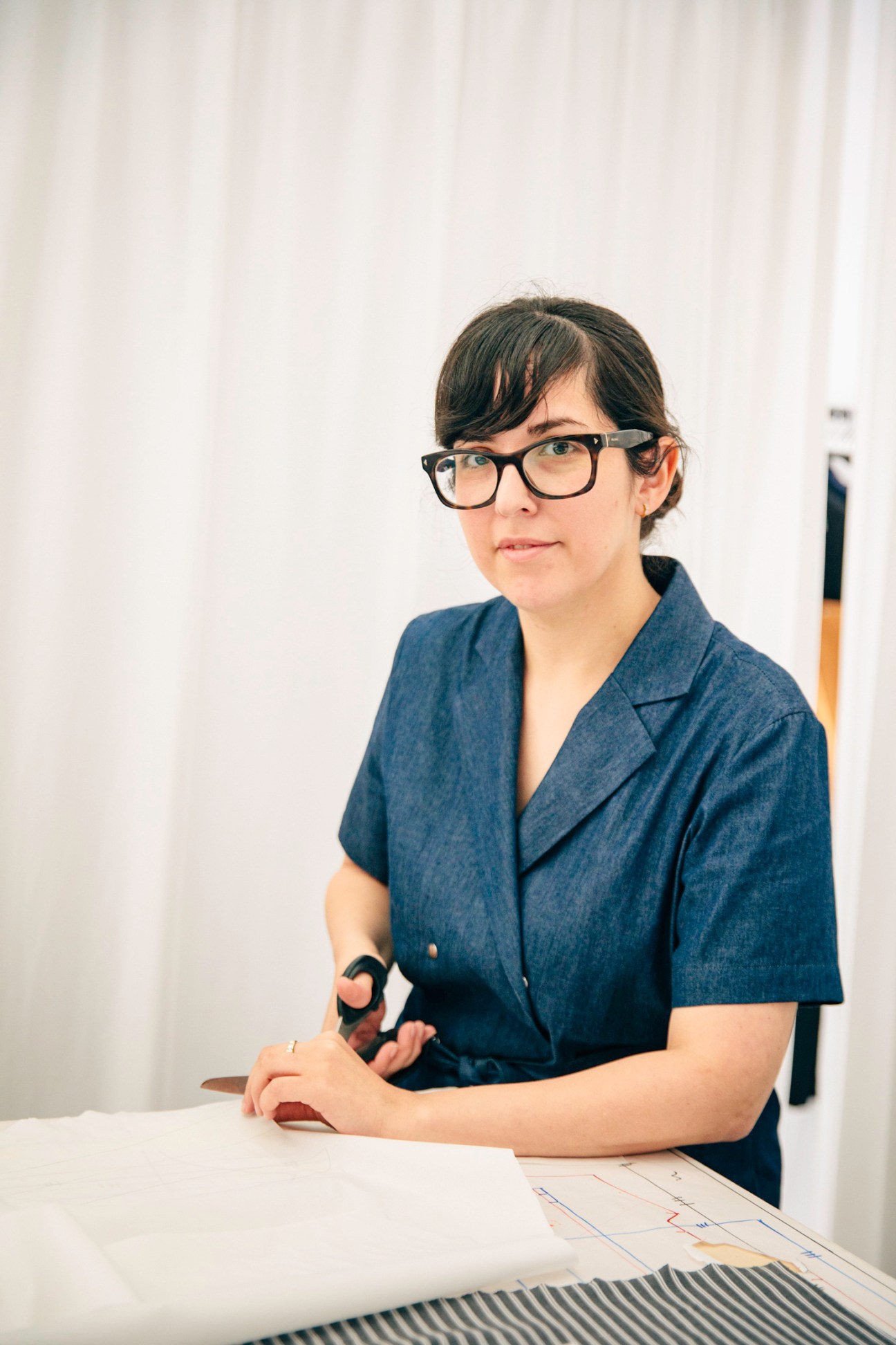 Woman wearing glasses and dark blue blazer sitting at table with design sketches.