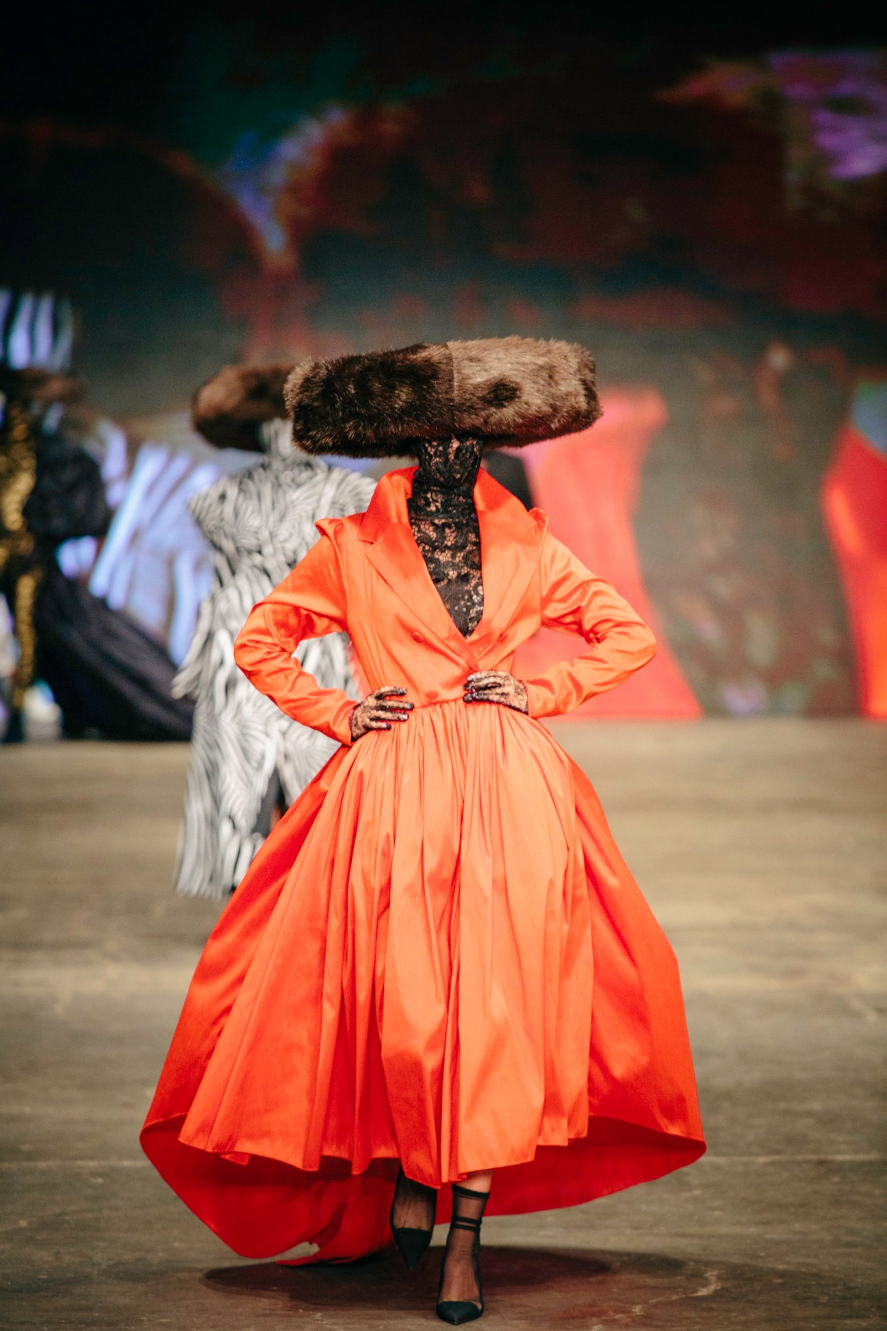 Model on runway wearing orange voluminous dress with large furry hat and lace collar.