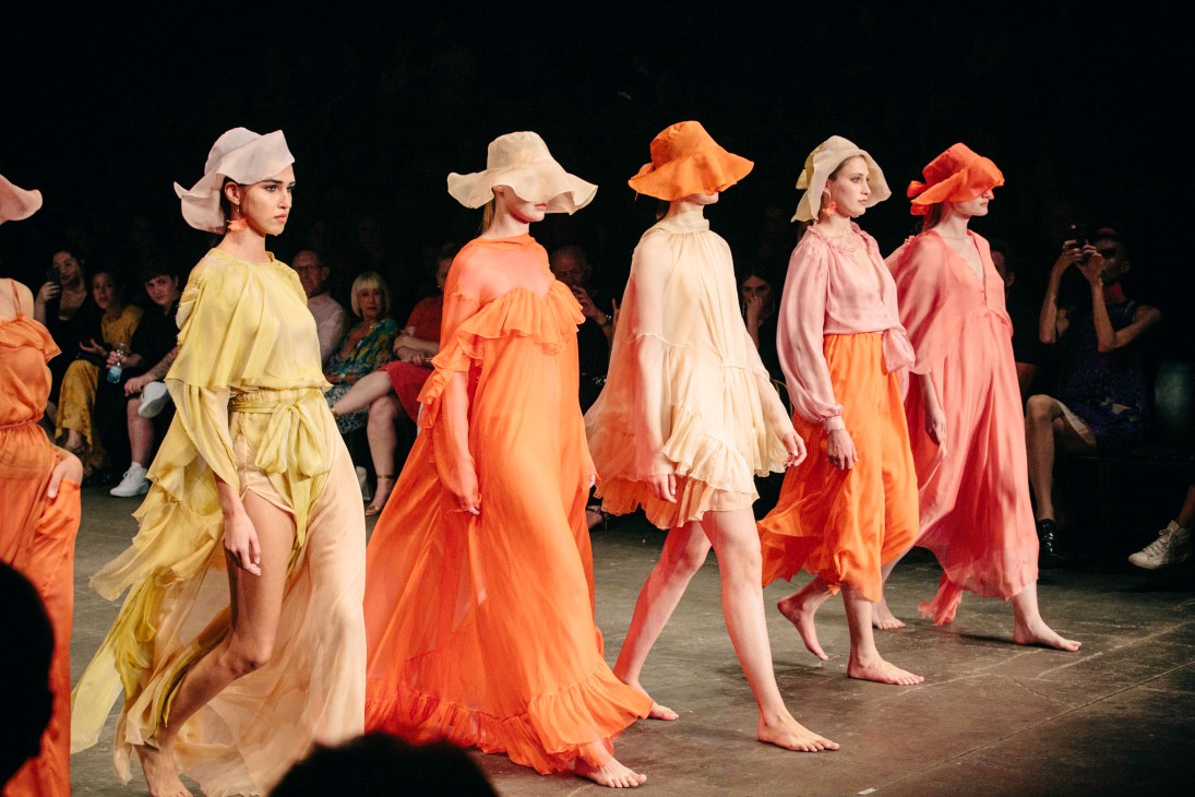 Models walk the runway wearing colorful flowing dresses in yellow, orange, cream, and pink with coordinating wide-brimmed hats from Nofar Refaeli's collection.