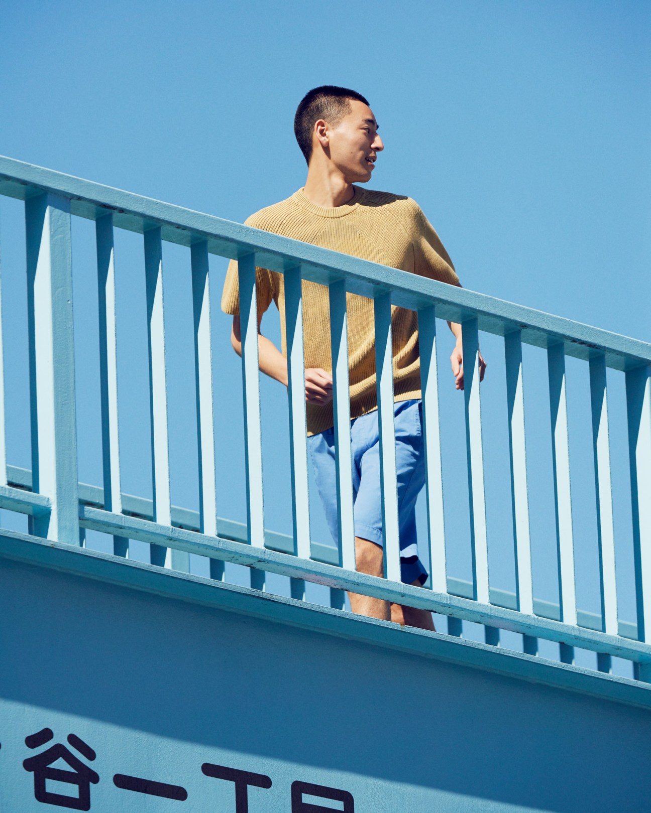 Man wearing a yellow ribbed jumper and blue shorts standing on a sky-blue pedestrian bridge.