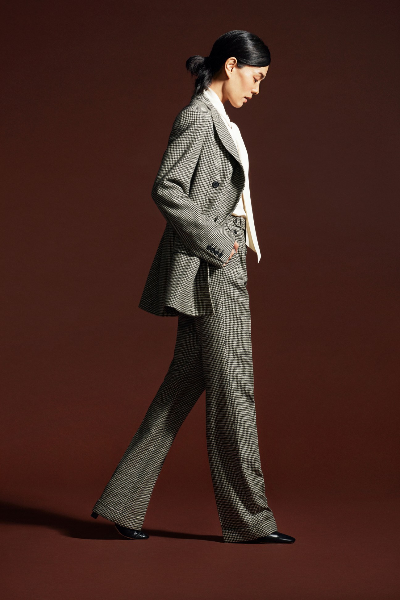 Woman in navy checked blazer and matching trousers with white bow blouse and black shoes against burgundy background