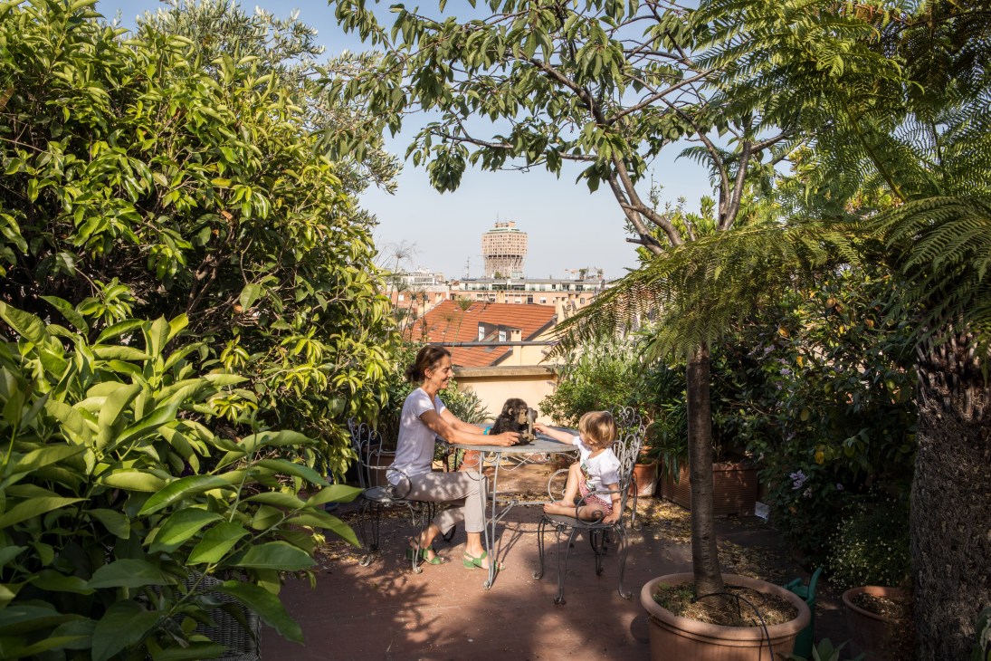 Woman and child sit at a table on a lush rooftop terrace surrounded by green plants and trees overlooking Milan's skyline.