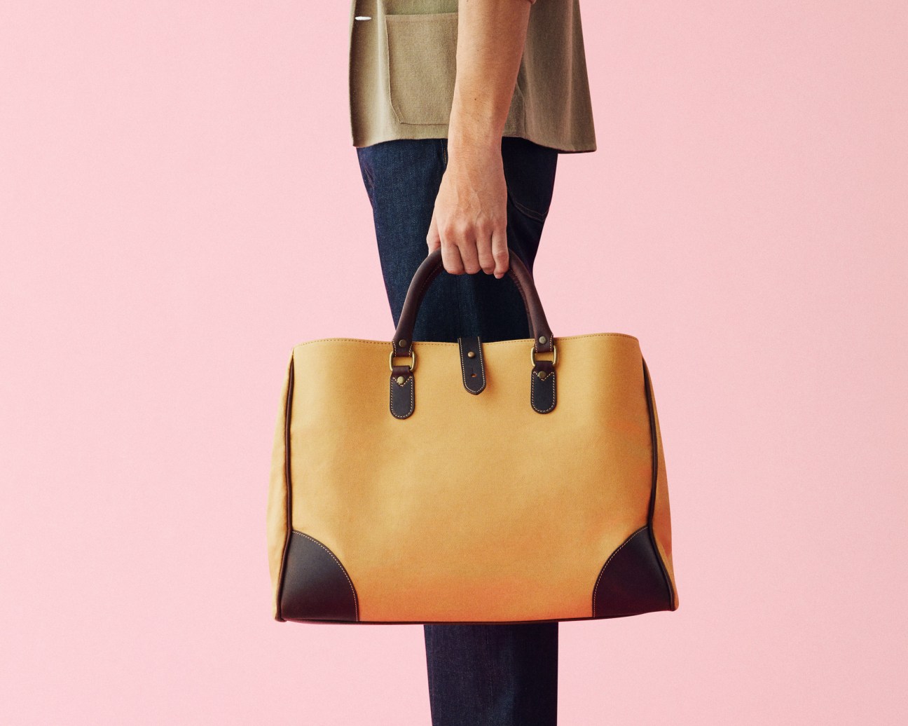 Person wearing a tan jacket and dark jeans holding a yellow and brown leather tote bag by Ettinger against a pink background.