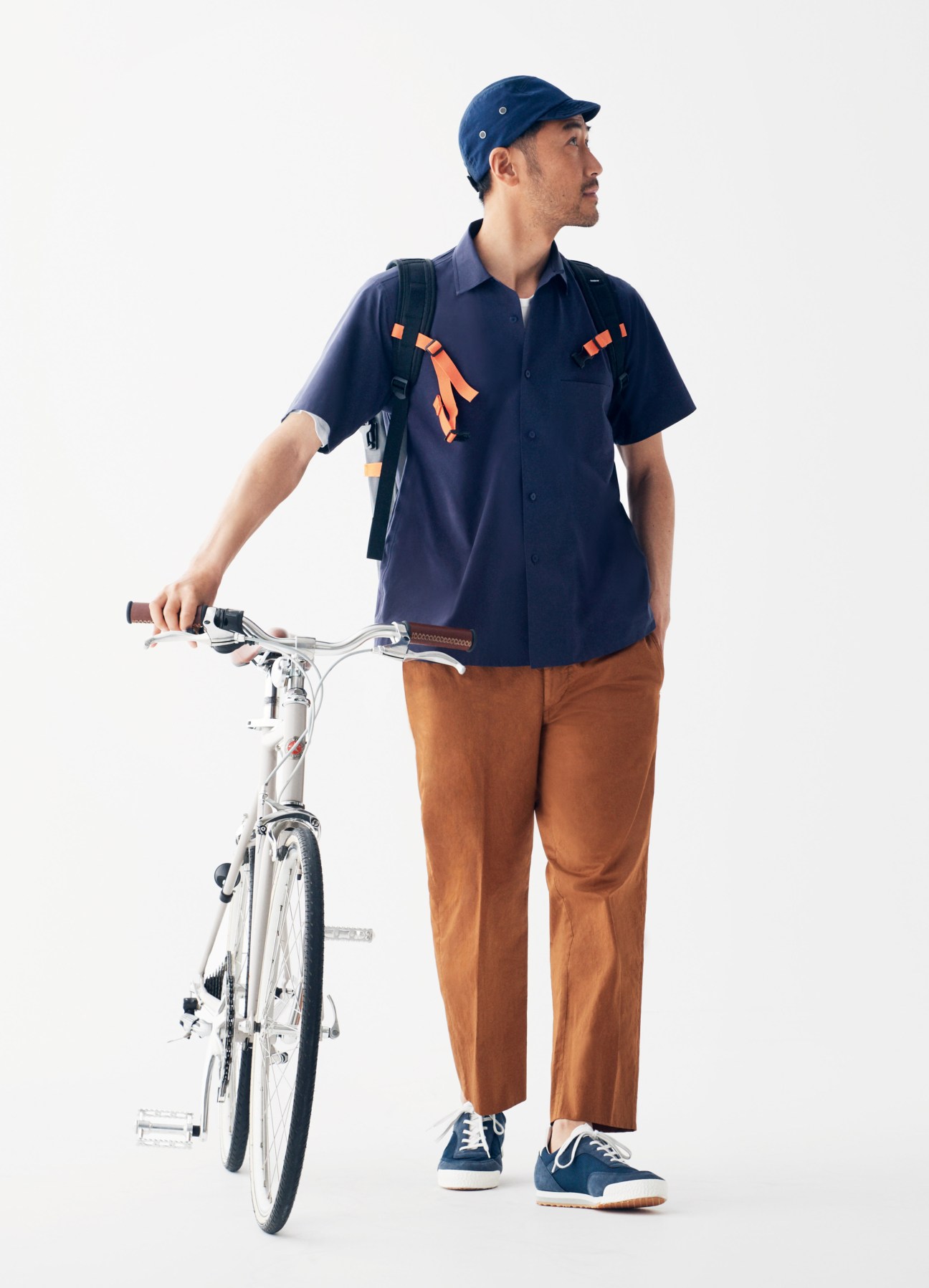 Man in navy shirt and brown trousers holding a white Tokyobike, wearing a blue cap and backpack.