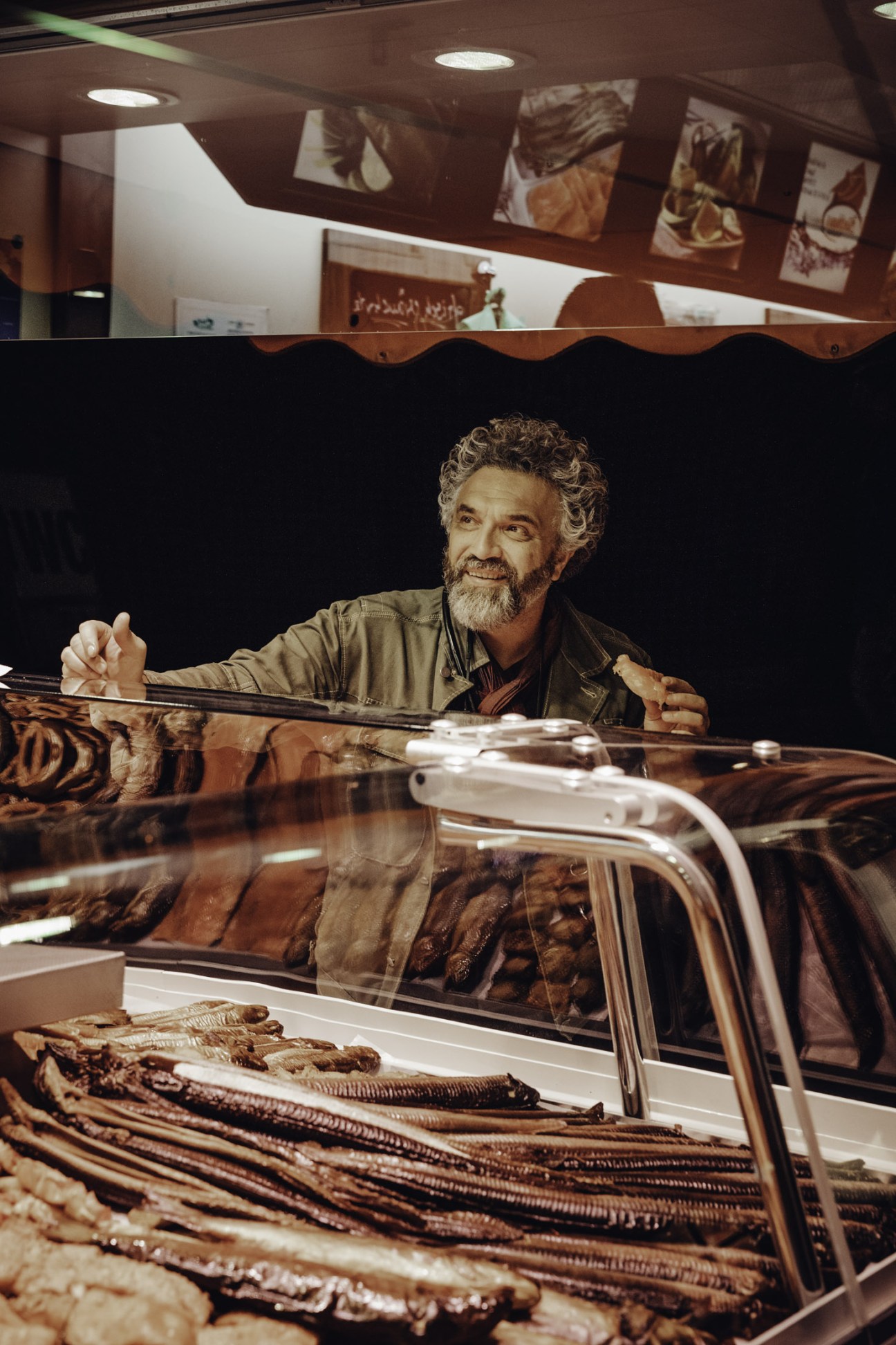 Man with curly grey hair and beard standing behind a fish market counter, smiling at customers.