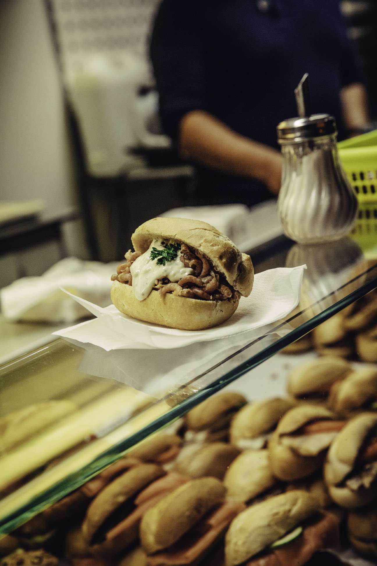 A fish sandwich with white sauce and herb garnish on a plate at a market counter.