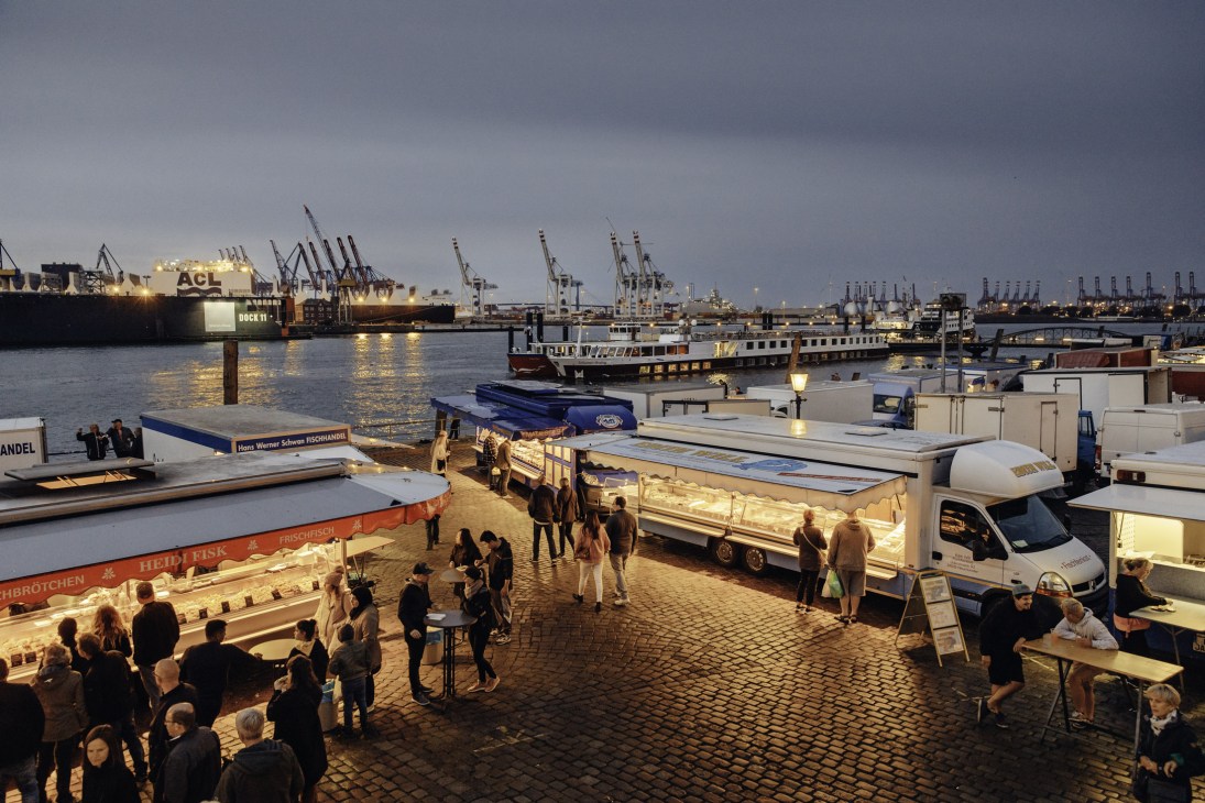 Early morning at Hamburg Fish Market with vendor stalls, boats, and shipping containers reflected in the Elbe River at sunrise.