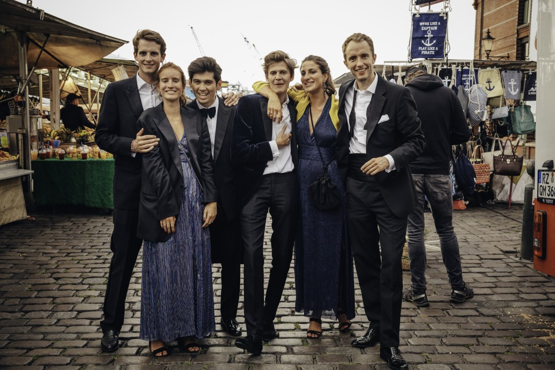 Six well-dressed wedding guests posing together at Hamburg's Fish Market on a cobblestone square.