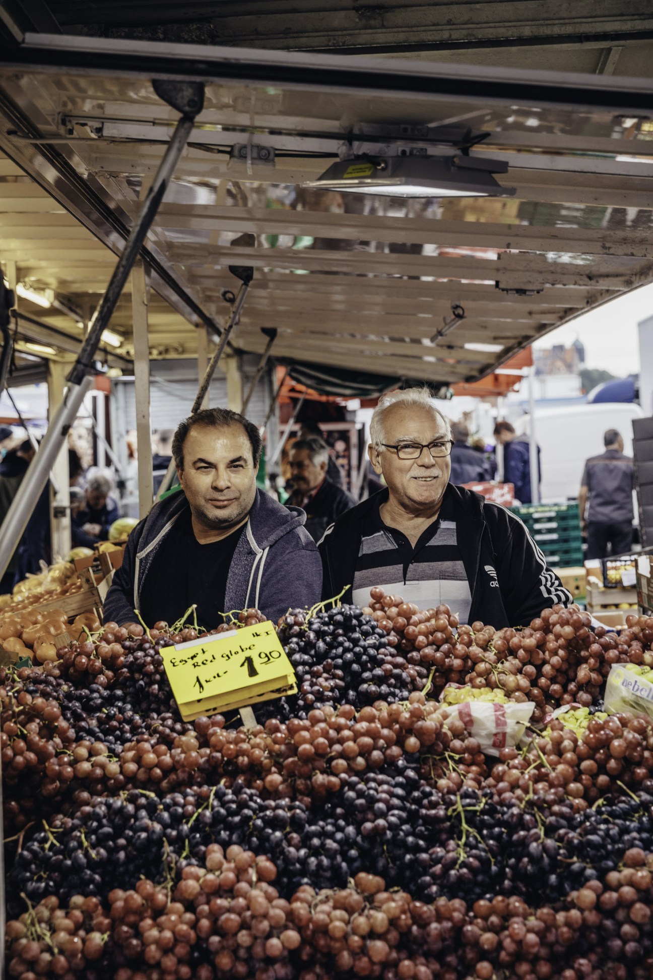 Two men stand behind a market stall displaying bunches of grapes in various colors with a yellow price sign.