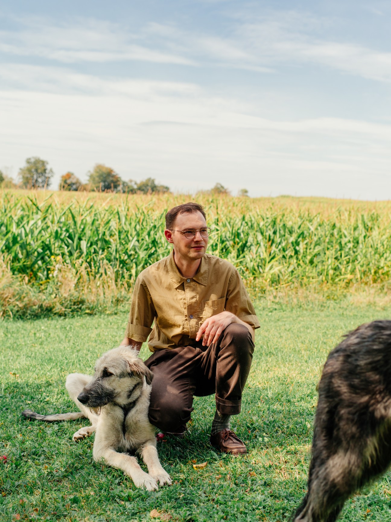 John Baker crouching in a grassy field beside two dogs with corn stalks in the background