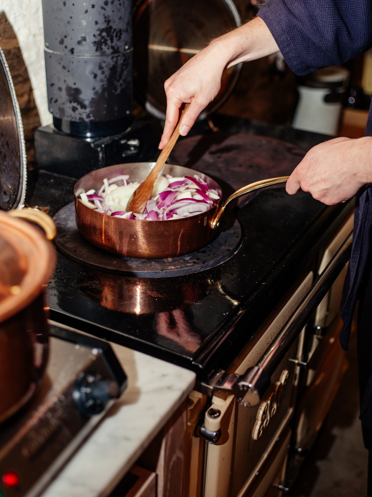 Person stirring sliced red and white onions in a copper pan with wooden spoon on stovetop