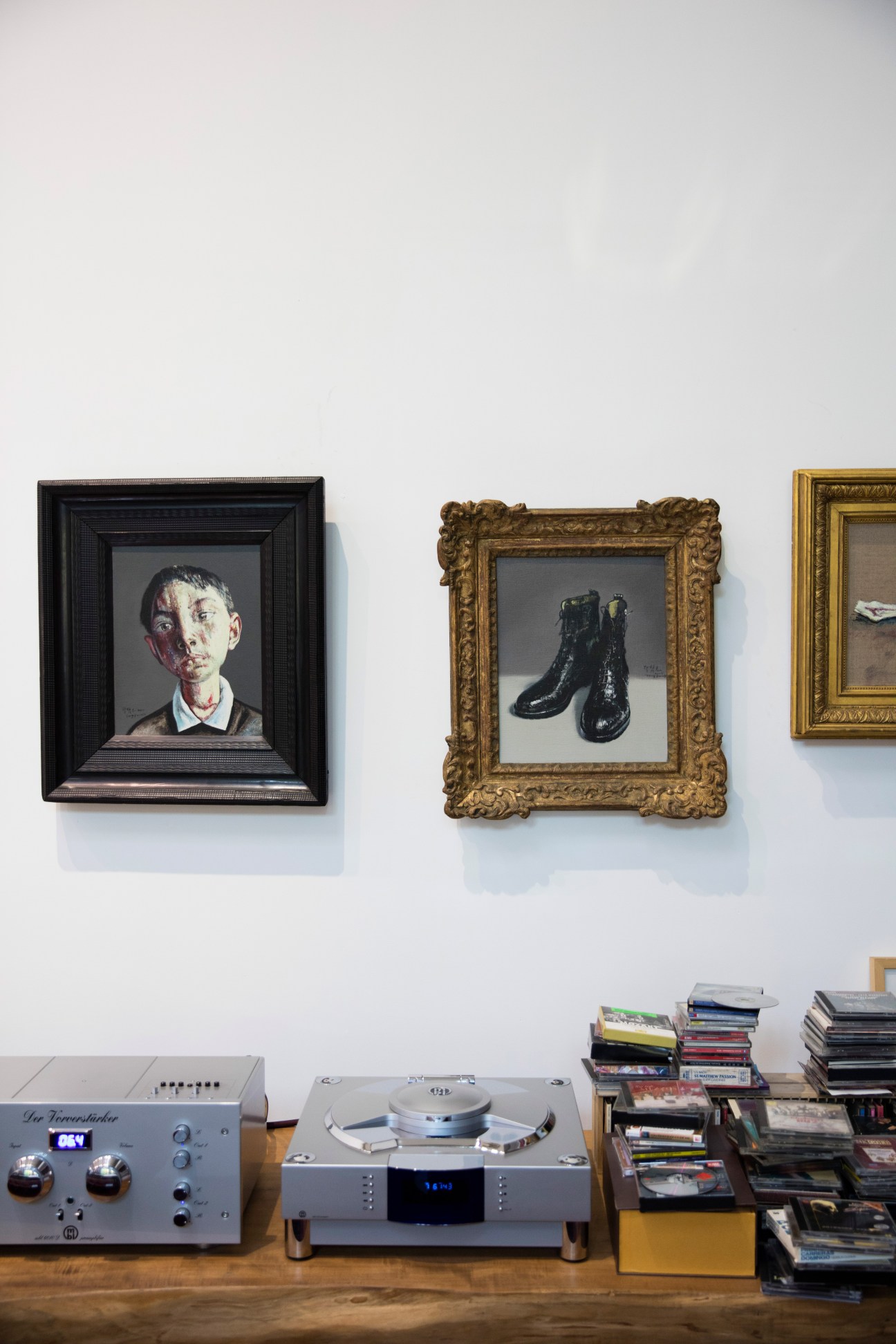 Studio wall displaying framed artworks including a portrait and boots, with audio equipment and stacked books below.