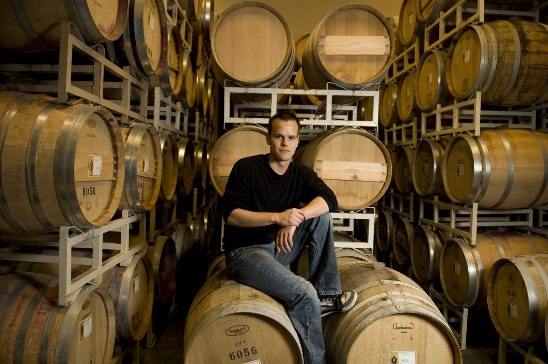 Aaron Crey, cellar assistant, sits on a wooden barrel surrounded by oak wine barrels at Nk'Mip Cellars.