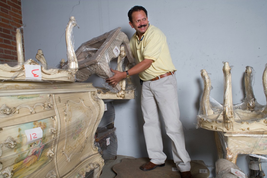 Man in yellow shirt examining ornate gilded furniture in a warehouse storage facility.