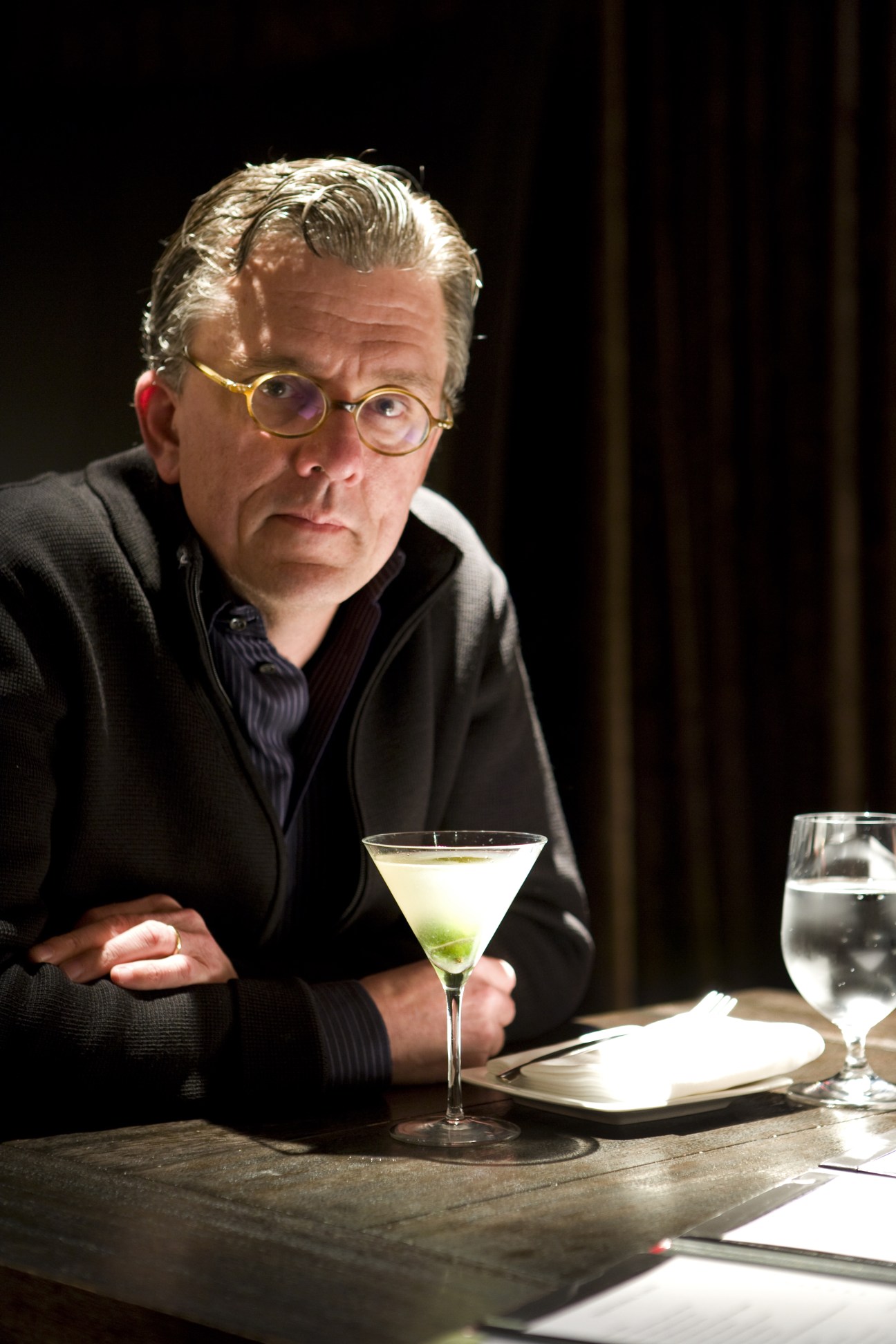 Kurt Andersen seated at a table holding a gin gimlet cocktail, wearing glasses and a dark jacket.