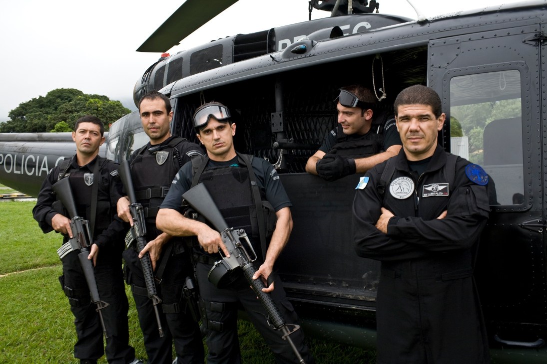Five armed police officers in tactical gear stand in front of a black UH-1H helicopter on a grass field.