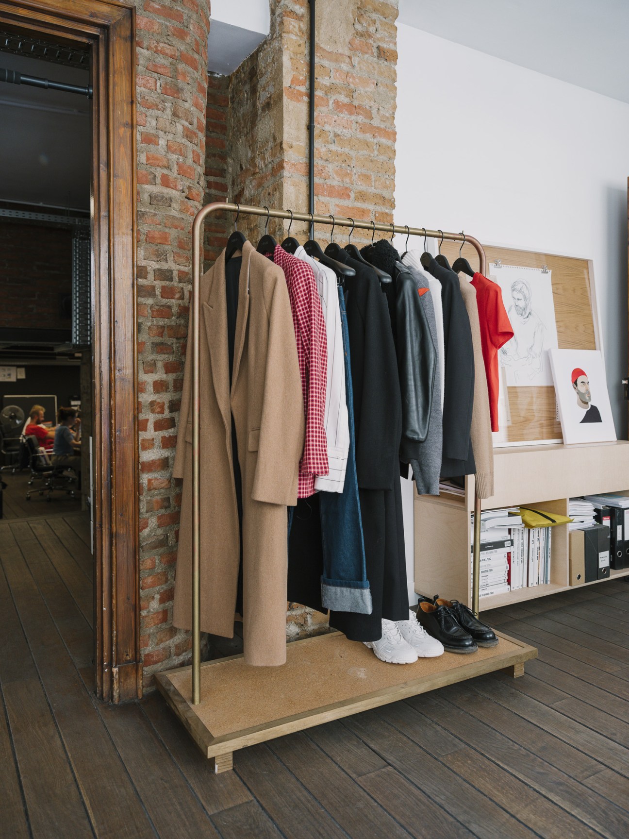 A brass clothing rail displaying Ami womenswear including coats, shirts, and a red beanie in an industrial studio space with exposed brick.