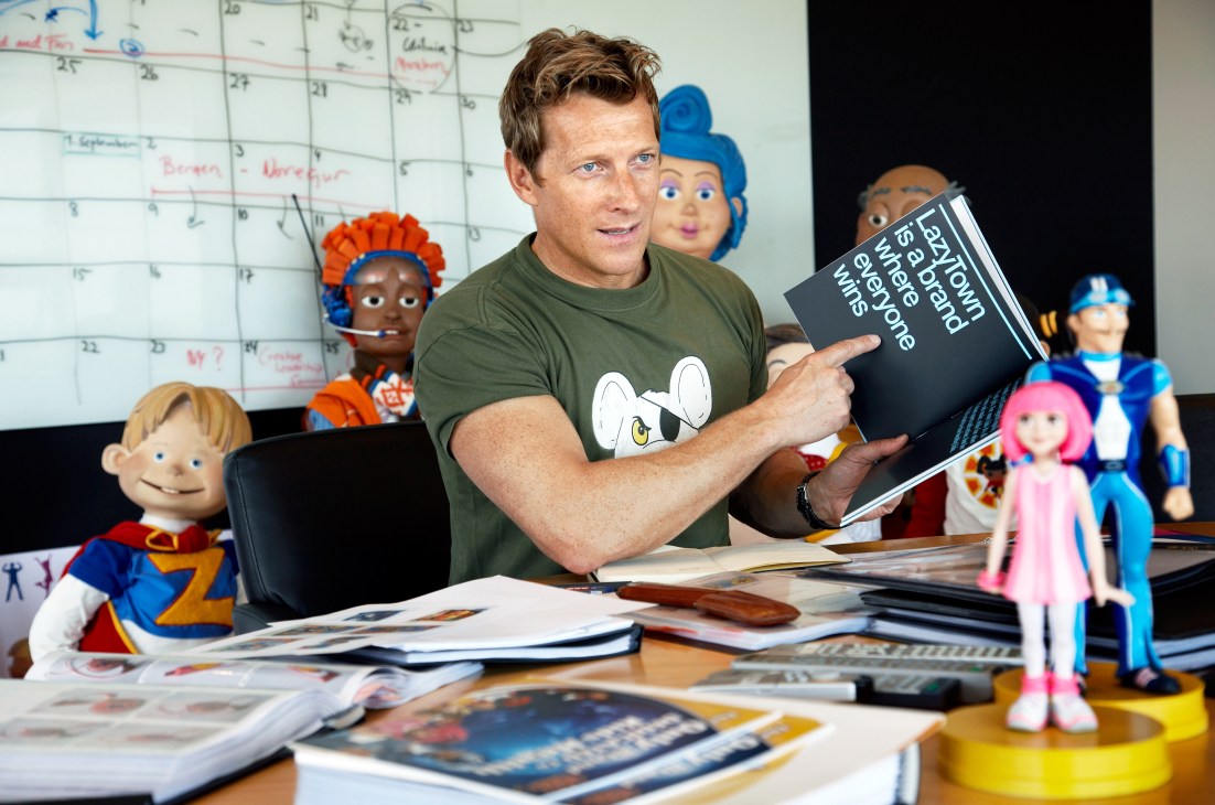 Magnus Scheving sits at a desk holding a LazyTown book, surrounded by colorful puppet characters in a studio.