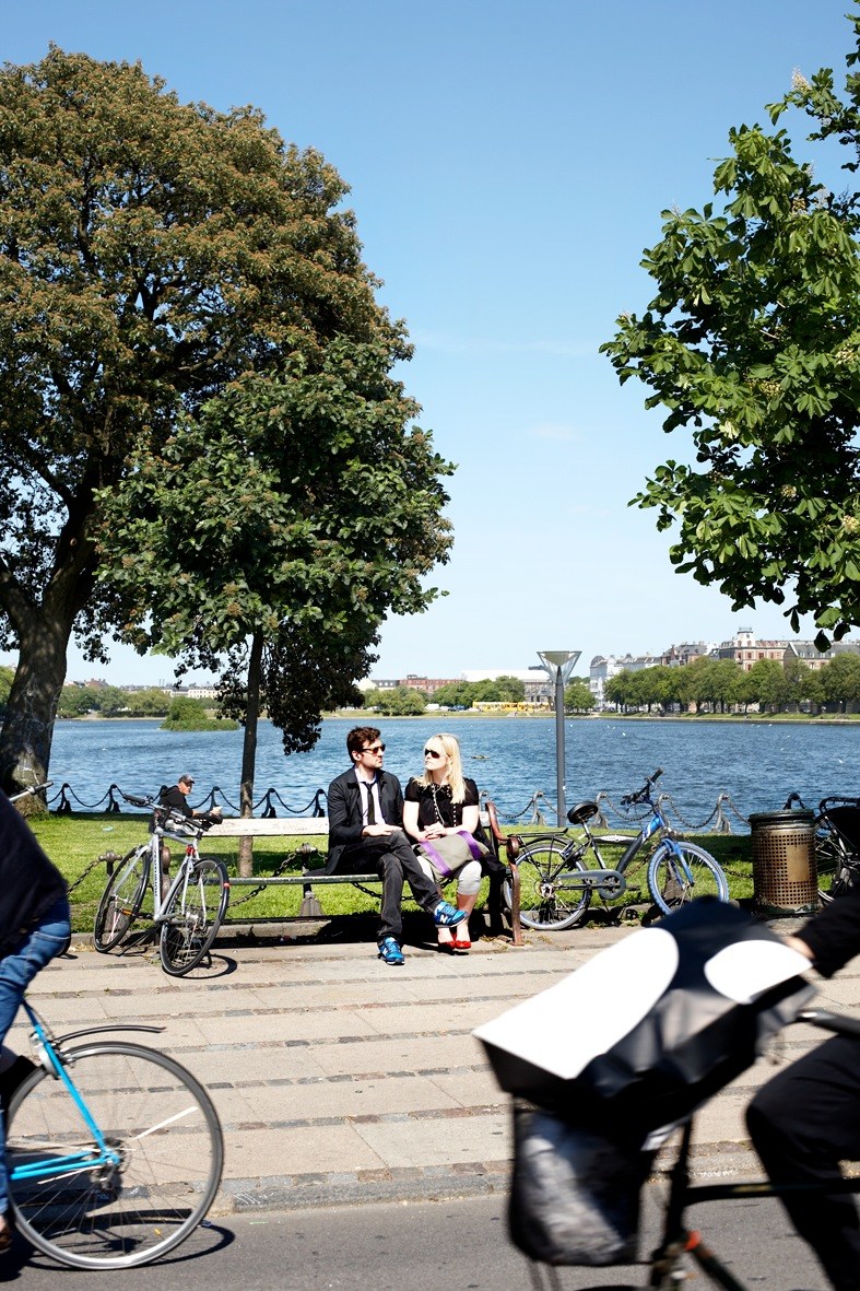Two people sitting on a bench by a waterfront with bicycles and trees nearby.