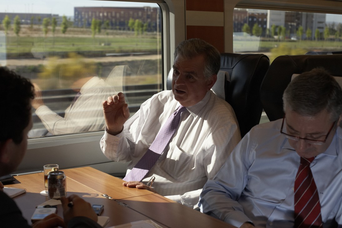 Ray LaHood gestures while speaking on a high-speed train with countryside visible through windows.