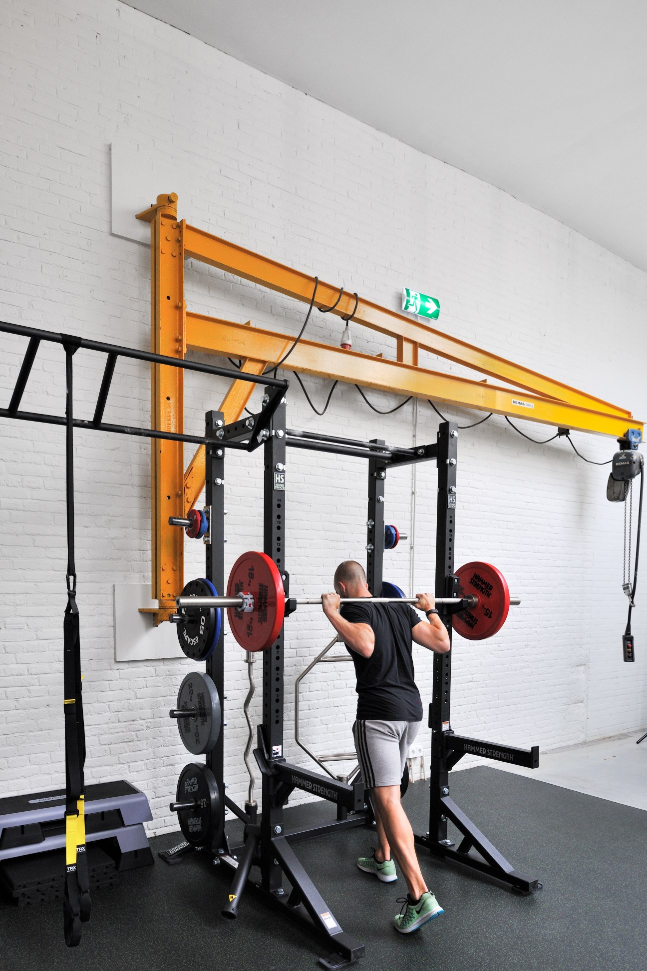 Man performing squats with barbell in office gym with orange crane beam overhead